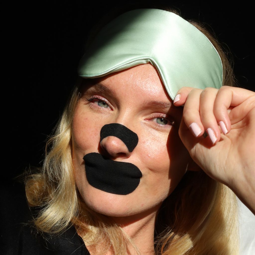 Vrouw met matzwarte neusstrip en mondtape kijkt in de camera en tilt haar slaapmasker op.