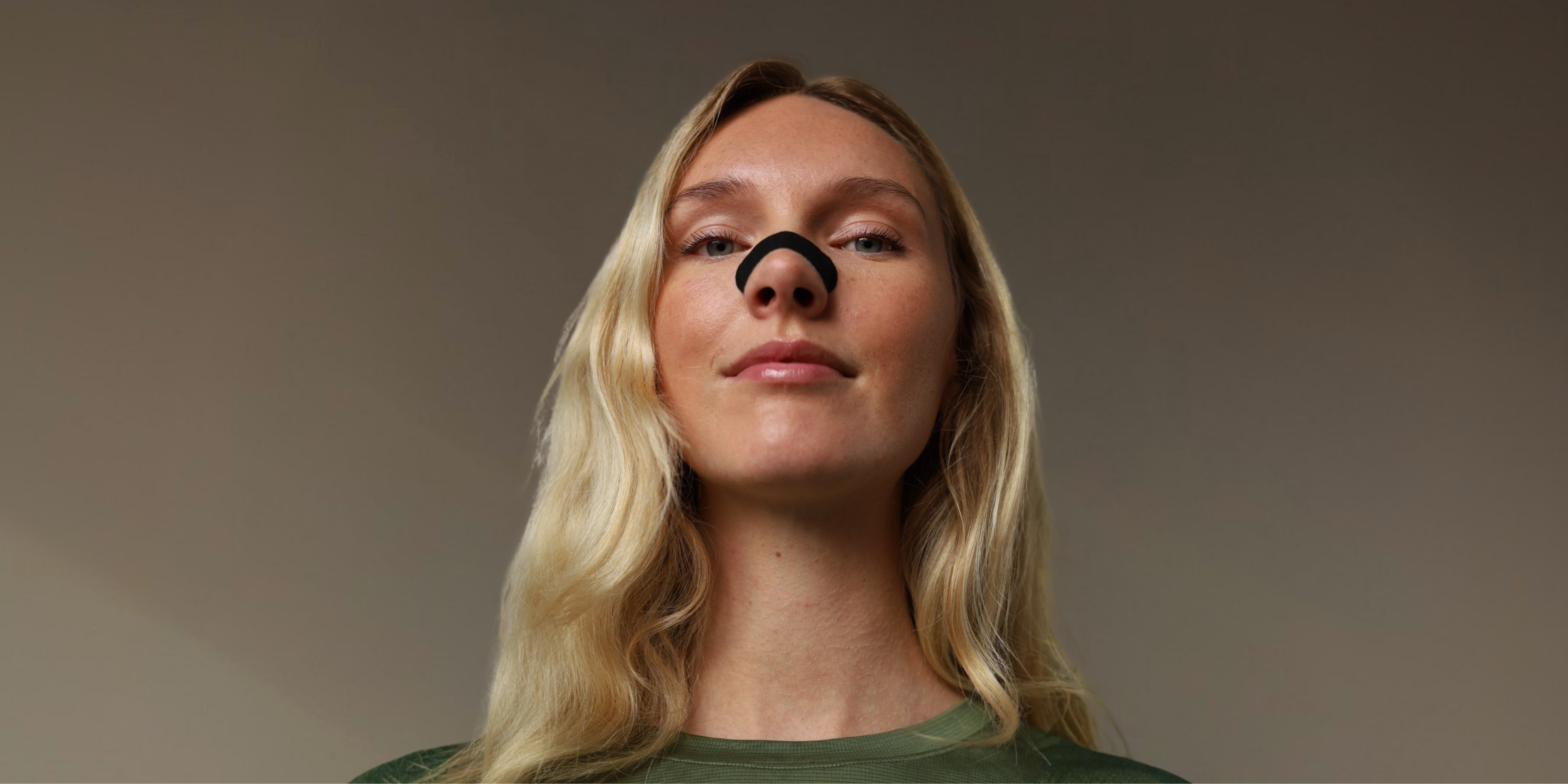 A low-angle, medium close-up photograph of a woman with long blonde hair, 25-35 years old, looking directly at the camera with a neutral expression against a solid taupe background. Positioned across her nose, is a black external nasal strip from The Sticky Mate™, demonstrating its application and how it physically holds her nasal passages more open for optimized nasal breathing.